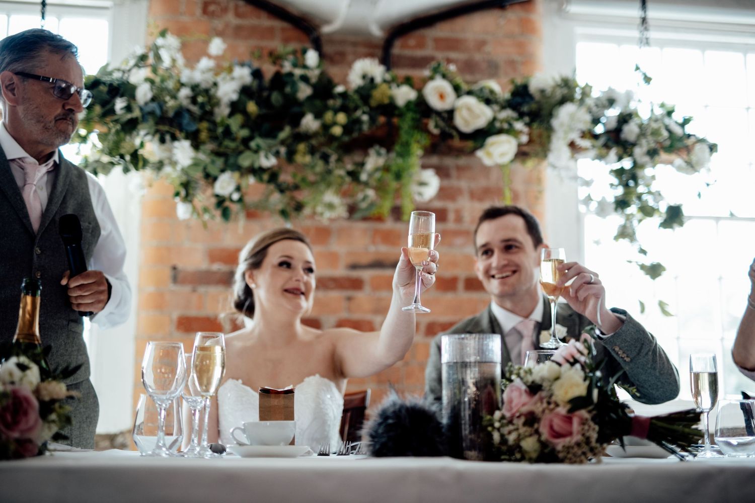 Wedding reception setup with floral arrangements and glassware in a rustic brick-walled venue.