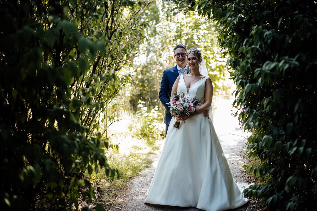Couple in wedding attire strolling through a sunlit garden pathway.