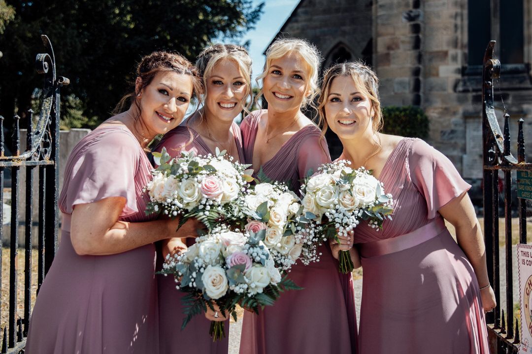 Wedding party members in mauve dresses pose together outdoors with white floral bouquets.