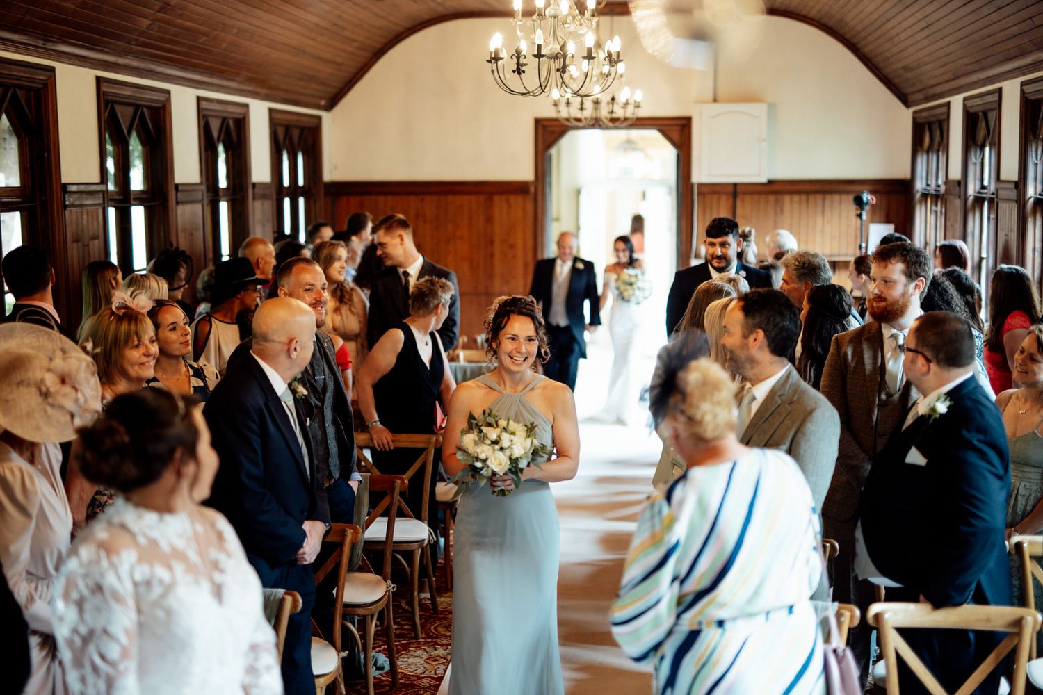 Wedding ceremony in a wooden-paneled room with chandeliers and rows of guests.
