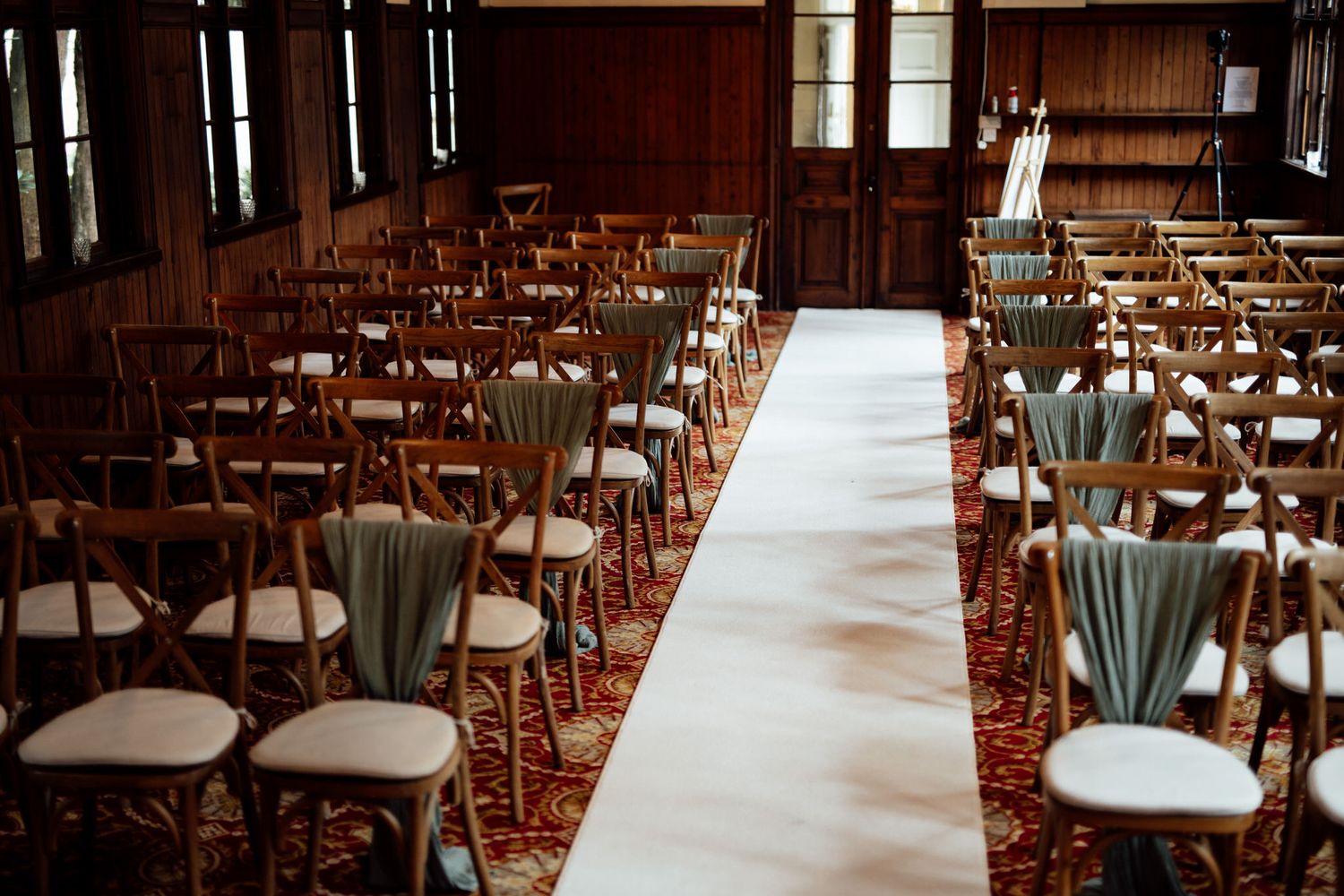 Elegant ceremony setup with white aisle runner and sage green chairs in a wood-paneled room.