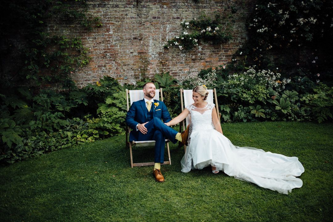 Wedding couple relaxing on lawn against brick wall with reception details.