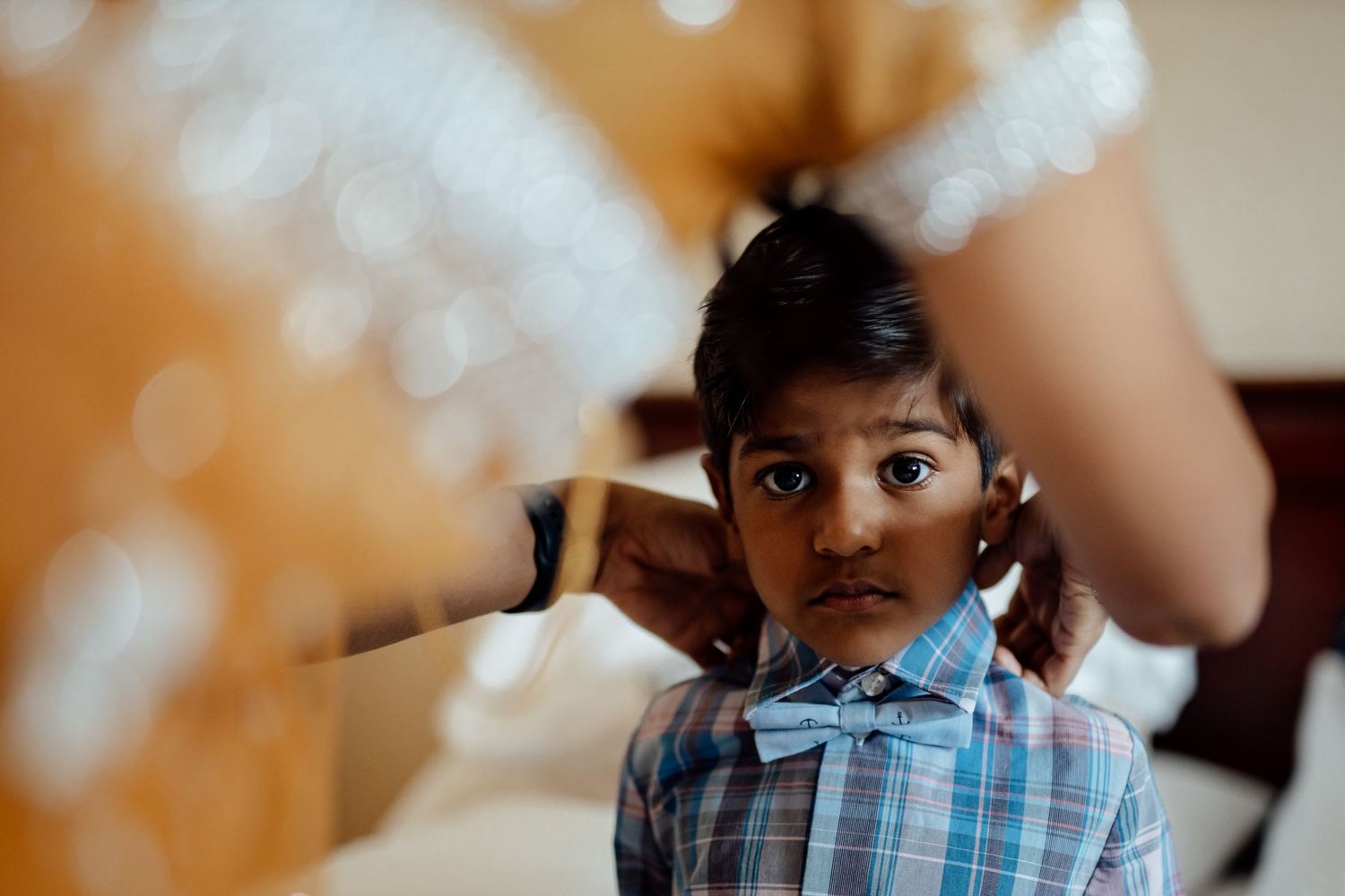 A young attendee in plaid bowtie peeks through wedding dress fabric.