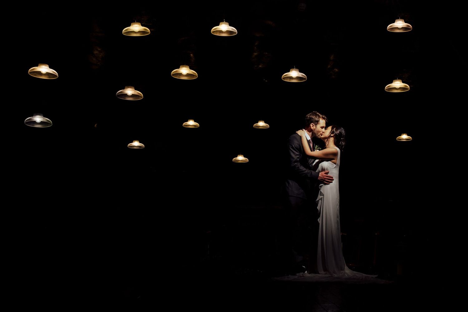 Romantic couple embracing under dramatic pendant lighting in dark room.