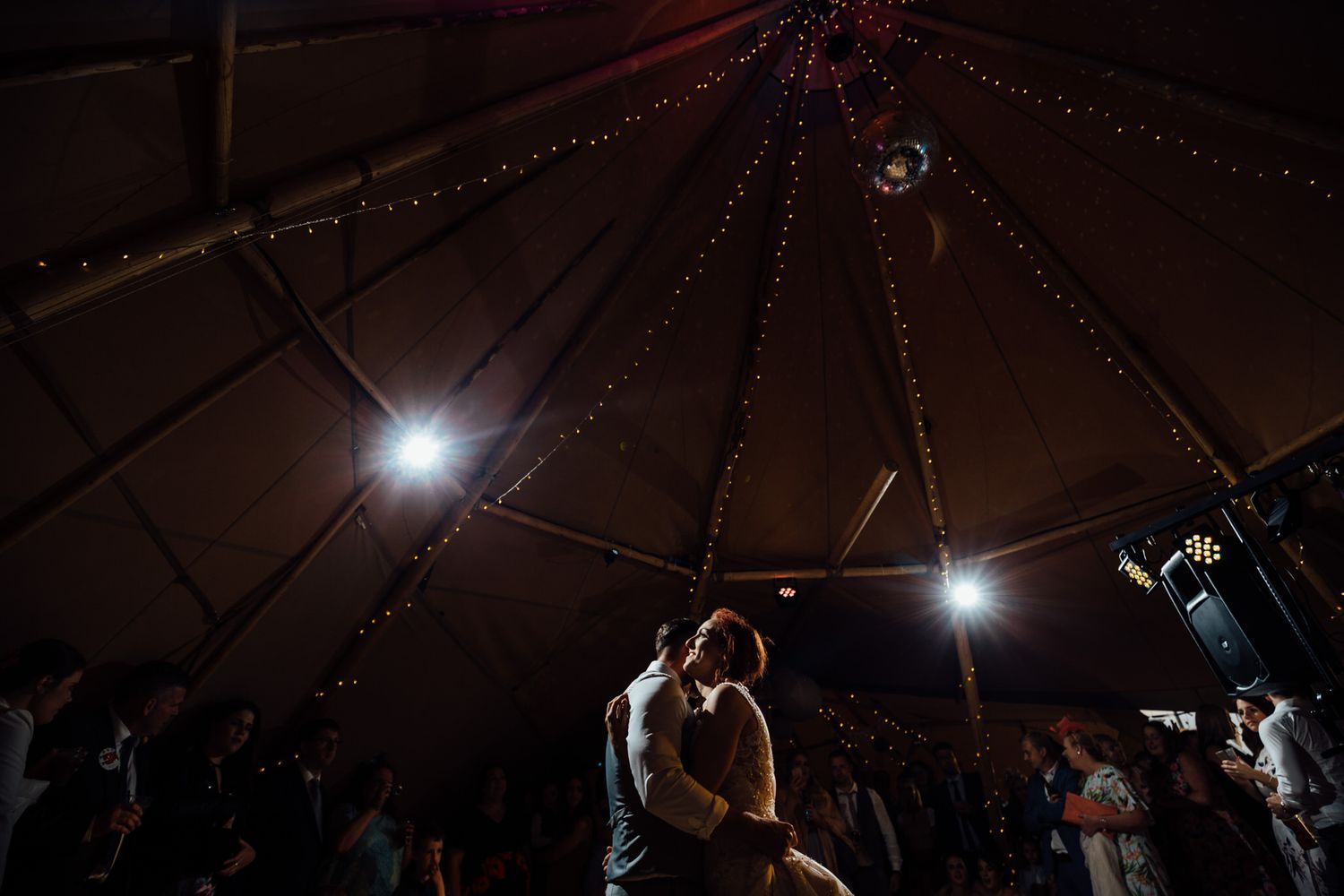 Dramatic lighting creates silhouettes of dancers in a dimly lit tipi wedding venue.
