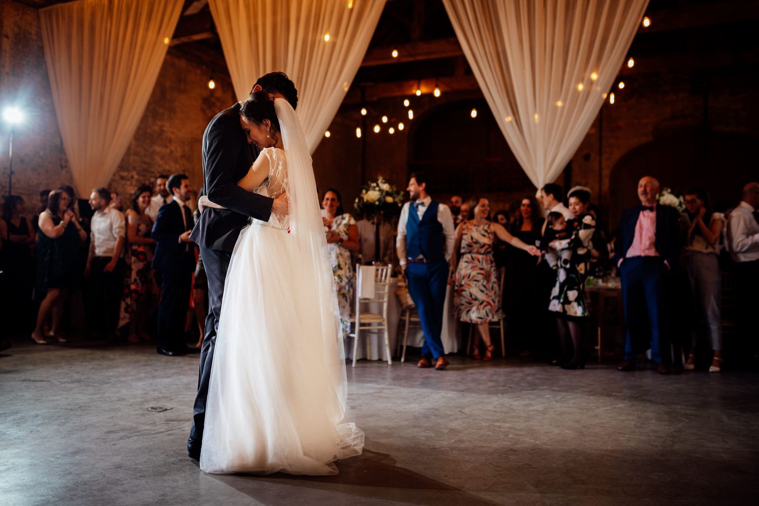Wedding reception guests dancing and celebrating on a dimly lit dance floor with white drapes and string lights.