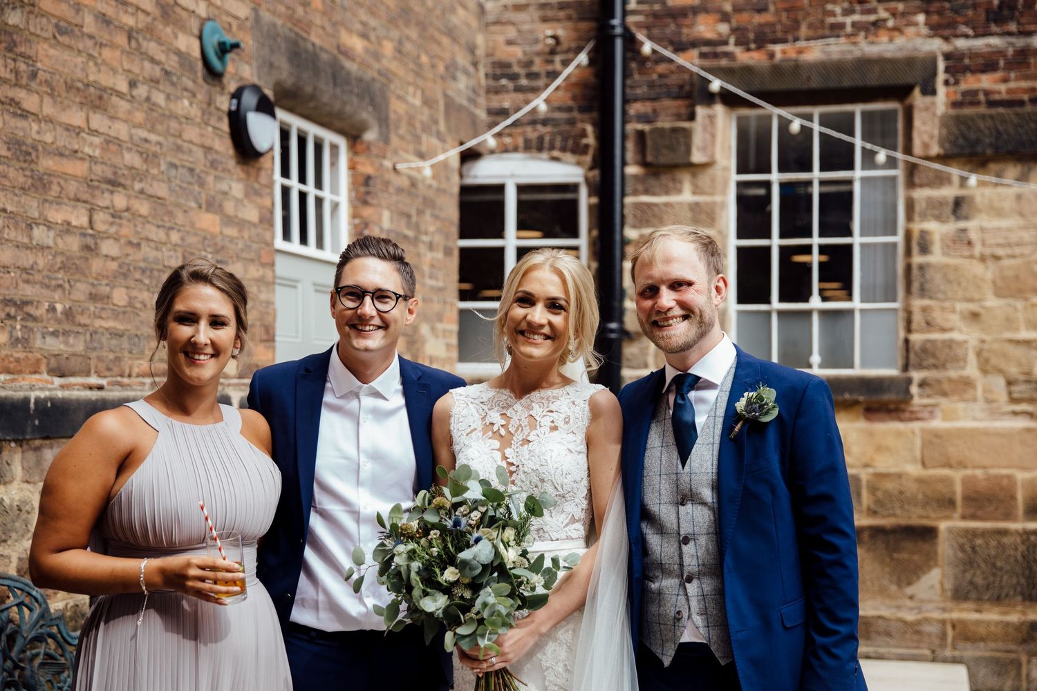 Wedding party posing against rustic brick wall in formal attire with floral bouquets.