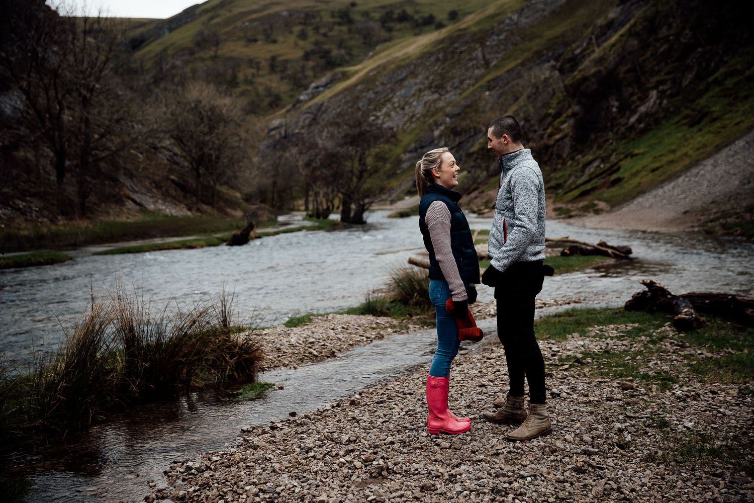 A couple stands together by a flowing river in a scenic mountain valley.