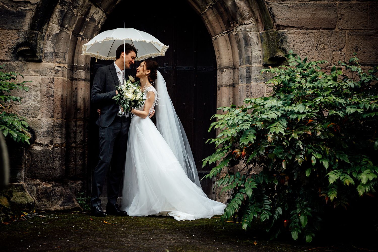 Wedding couple poses under stone archways surrounded by lush green foliage.