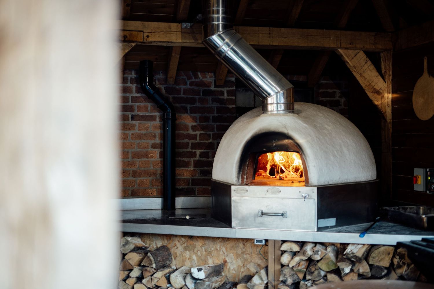 Wood-fired pizza oven with flames visible inside brick and metal structure.