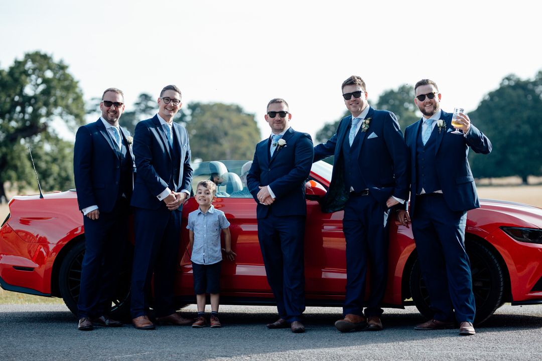Wedding party poses with classic red vintage car on sunny outdoor path.