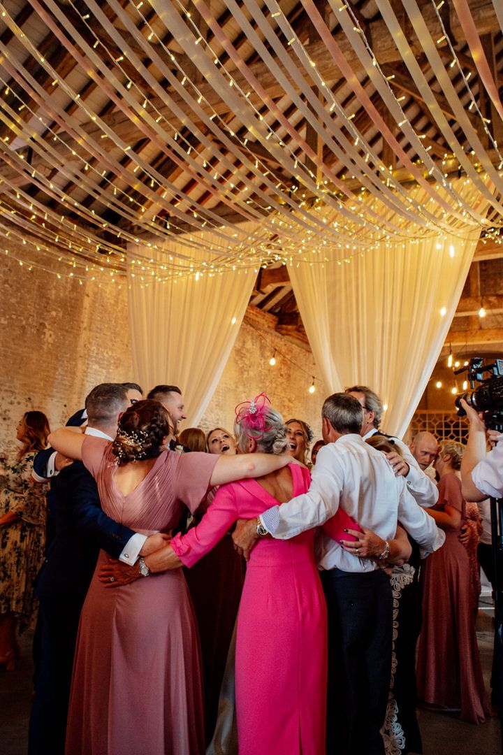 Group of wedding guests dance in a line under twinkling lights and white ceiling drapes.