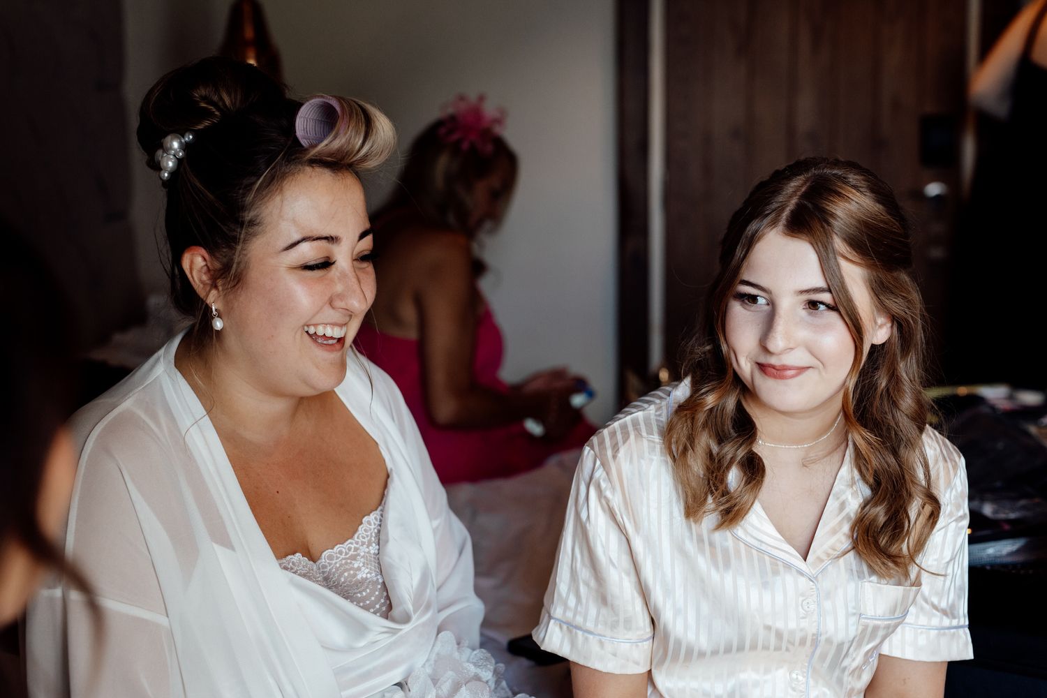 Two women in white attire share a candid moment during wedding preparations.