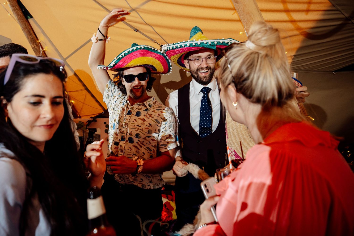 Party guests celebrating under tent with someone wearing colorful sombrero and festive attire.