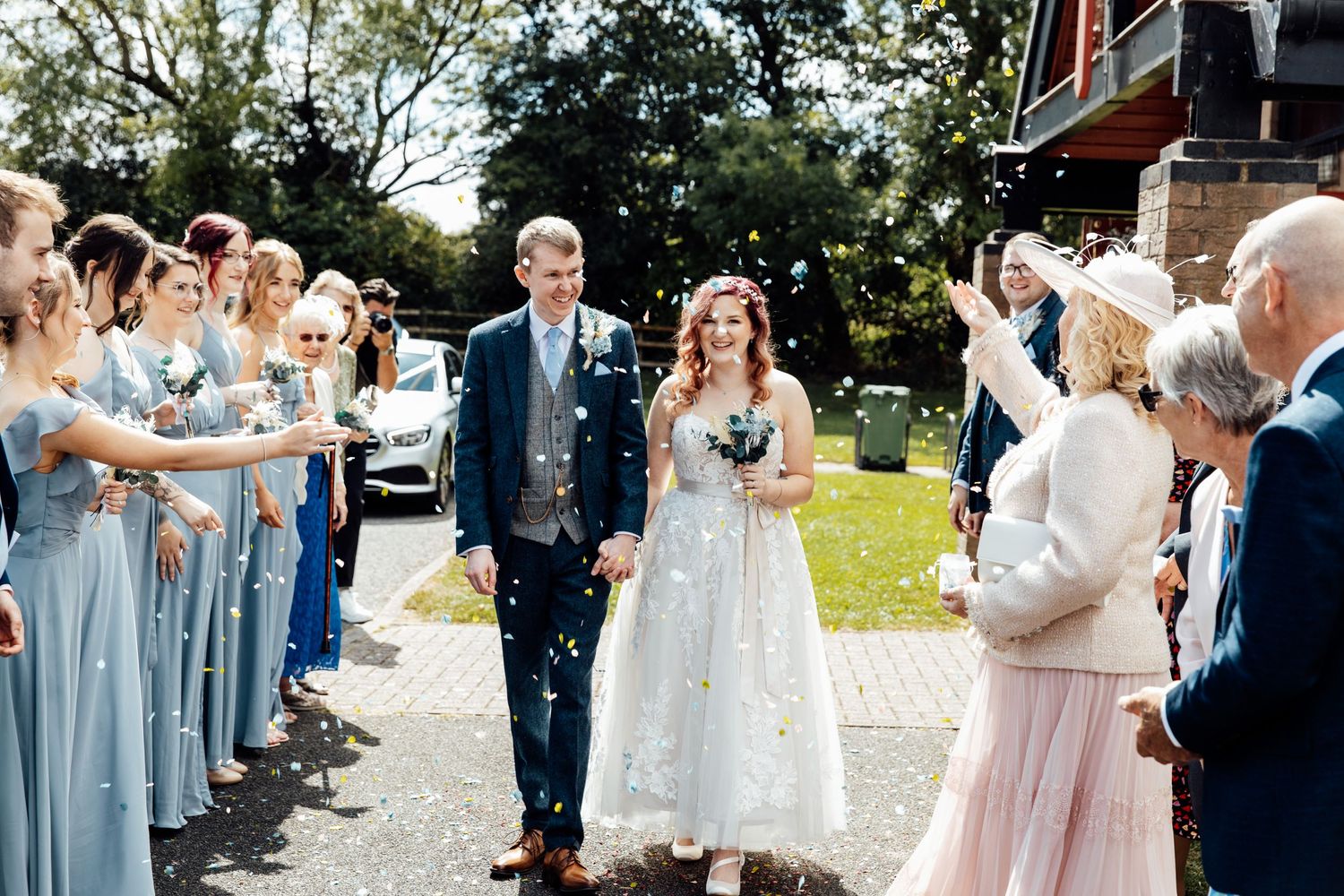 Wedding guests throw confetti as couple walks through celebration line outdoors.