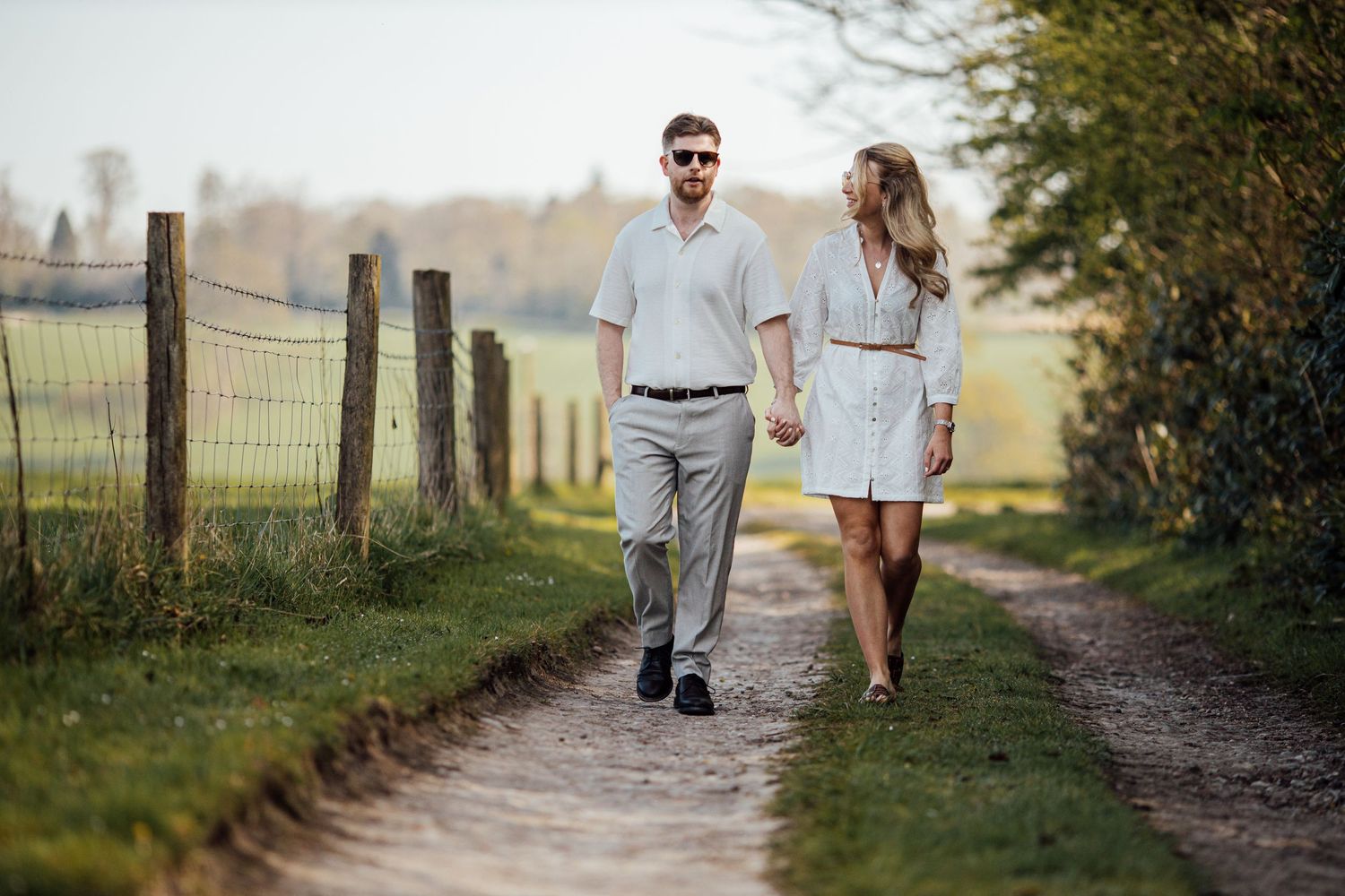 A couple in white outfits walking together along a scenic countryside path during golden hour.