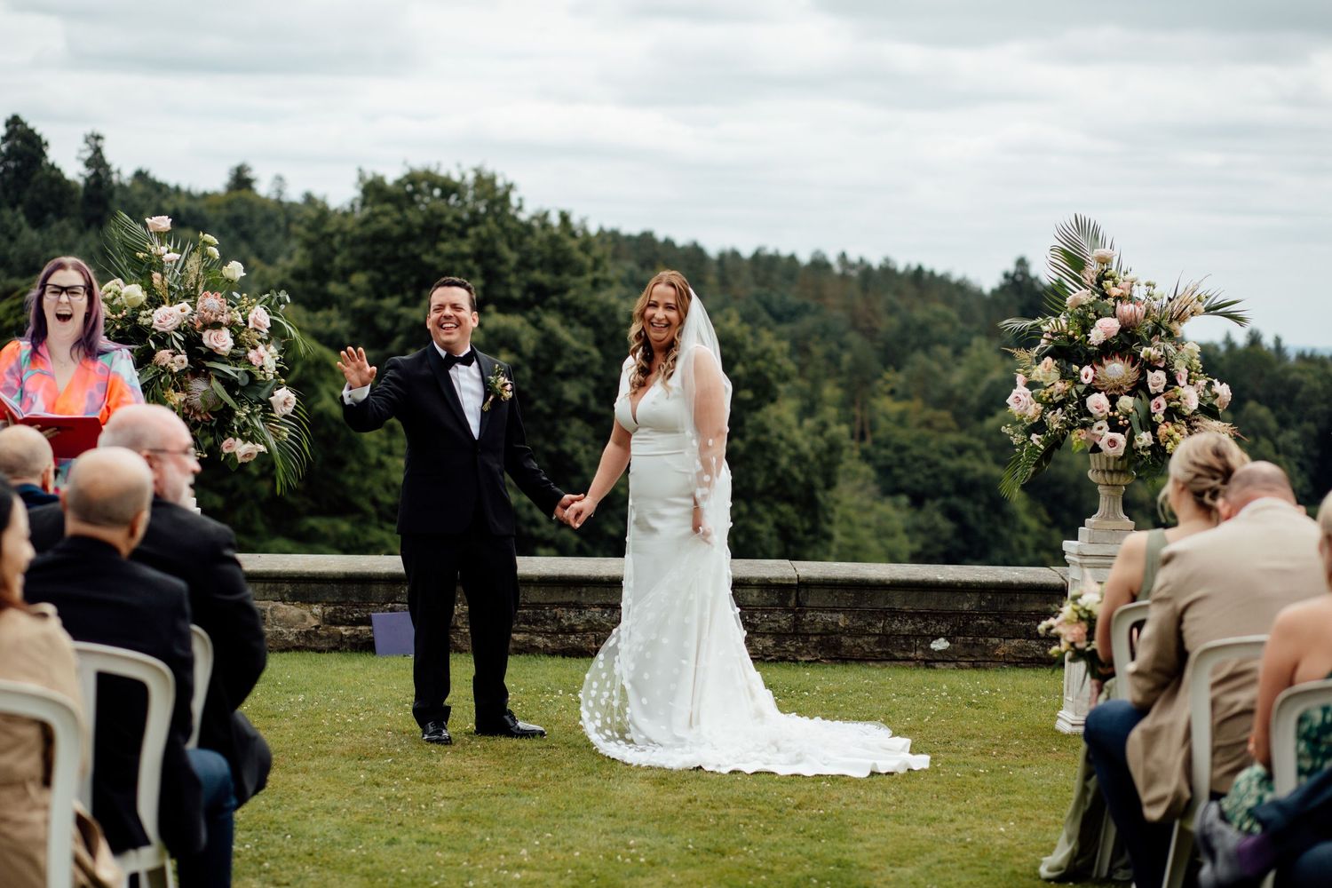 Outdoor wedding ceremony with floral arch arrangements and seated guests on lawn.