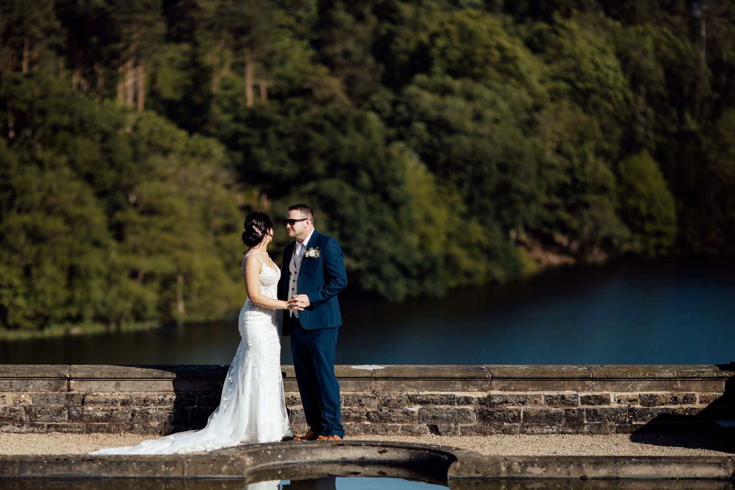 Wedding couple poses on stone bridge overlooking serene lake surrounded by forest.