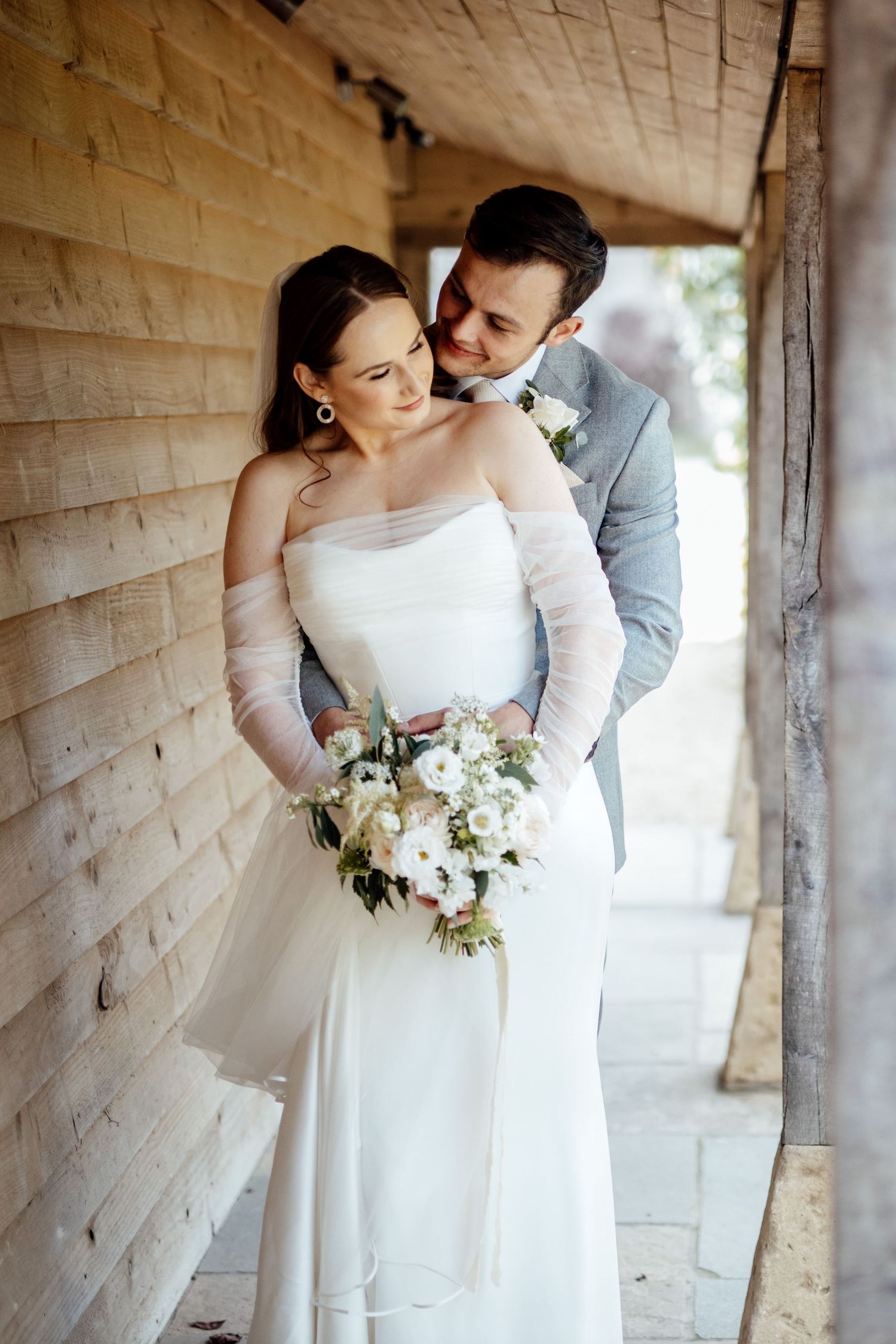 Romantic wedding portraits against a white brick wall with the couple holding a white floral bouquet.