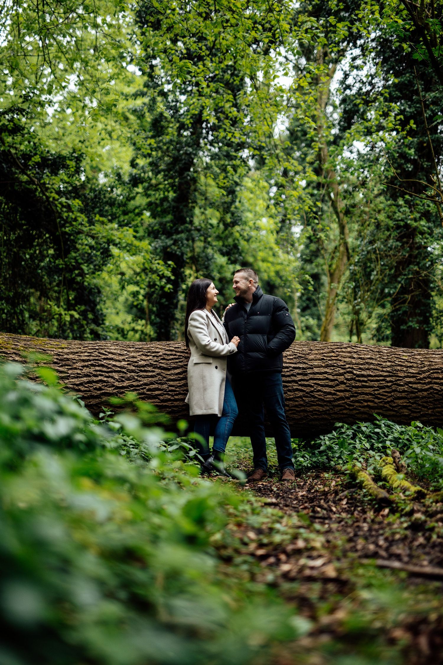Two people share an intimate moment while sitting on a large fallen tree trunk in a wooded area.
