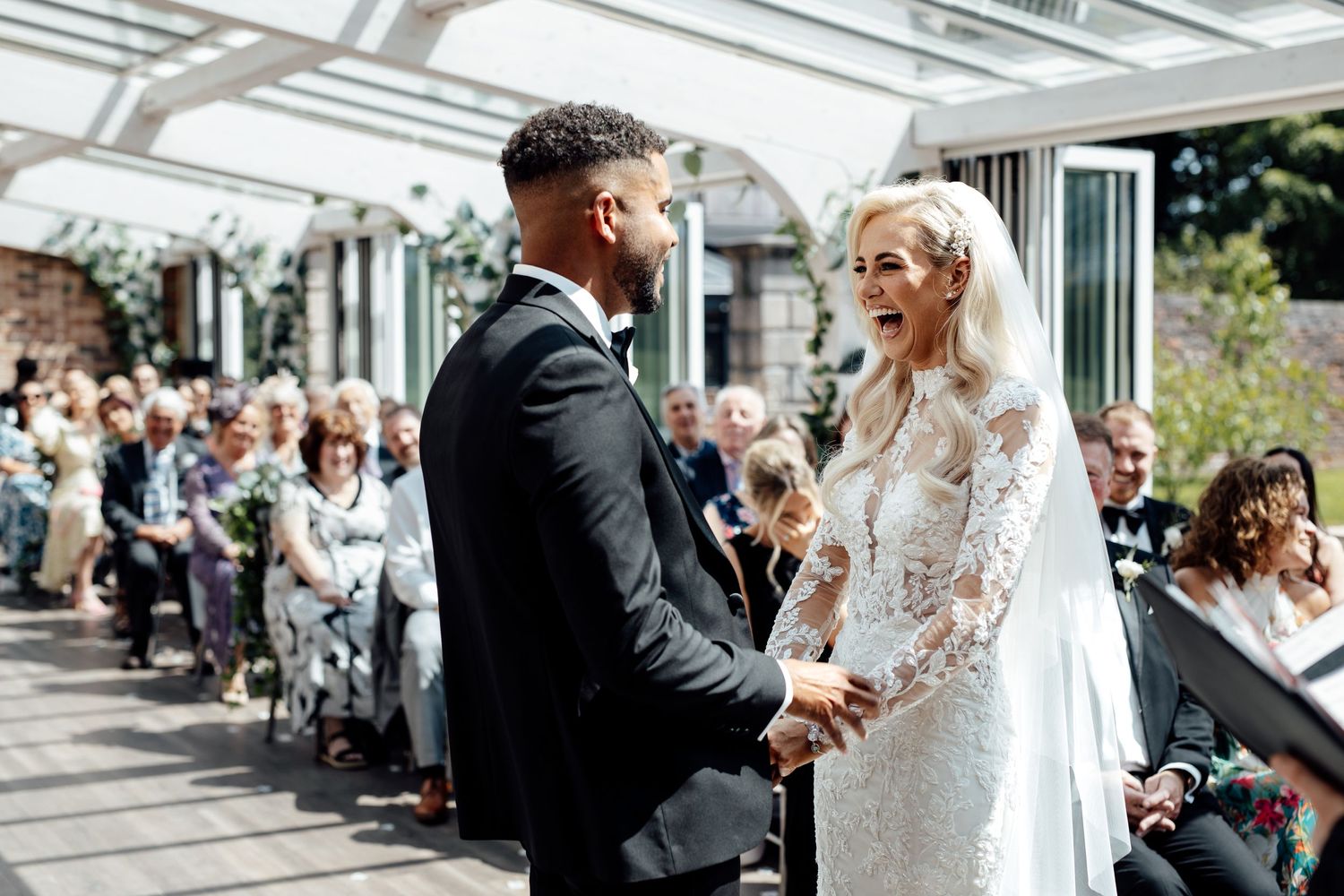 Elegant wedding ceremony with guests watching a couple exchange vows under a glass-roofed venue.