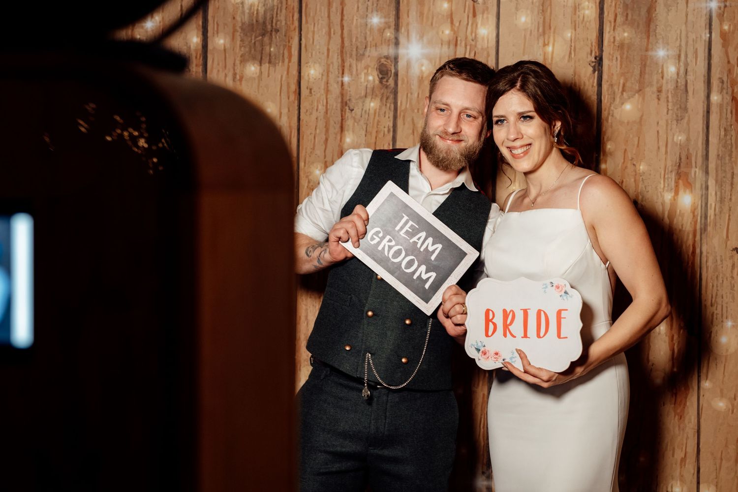 Couple poses with wedding signs against rustic wooden wall backdrop.