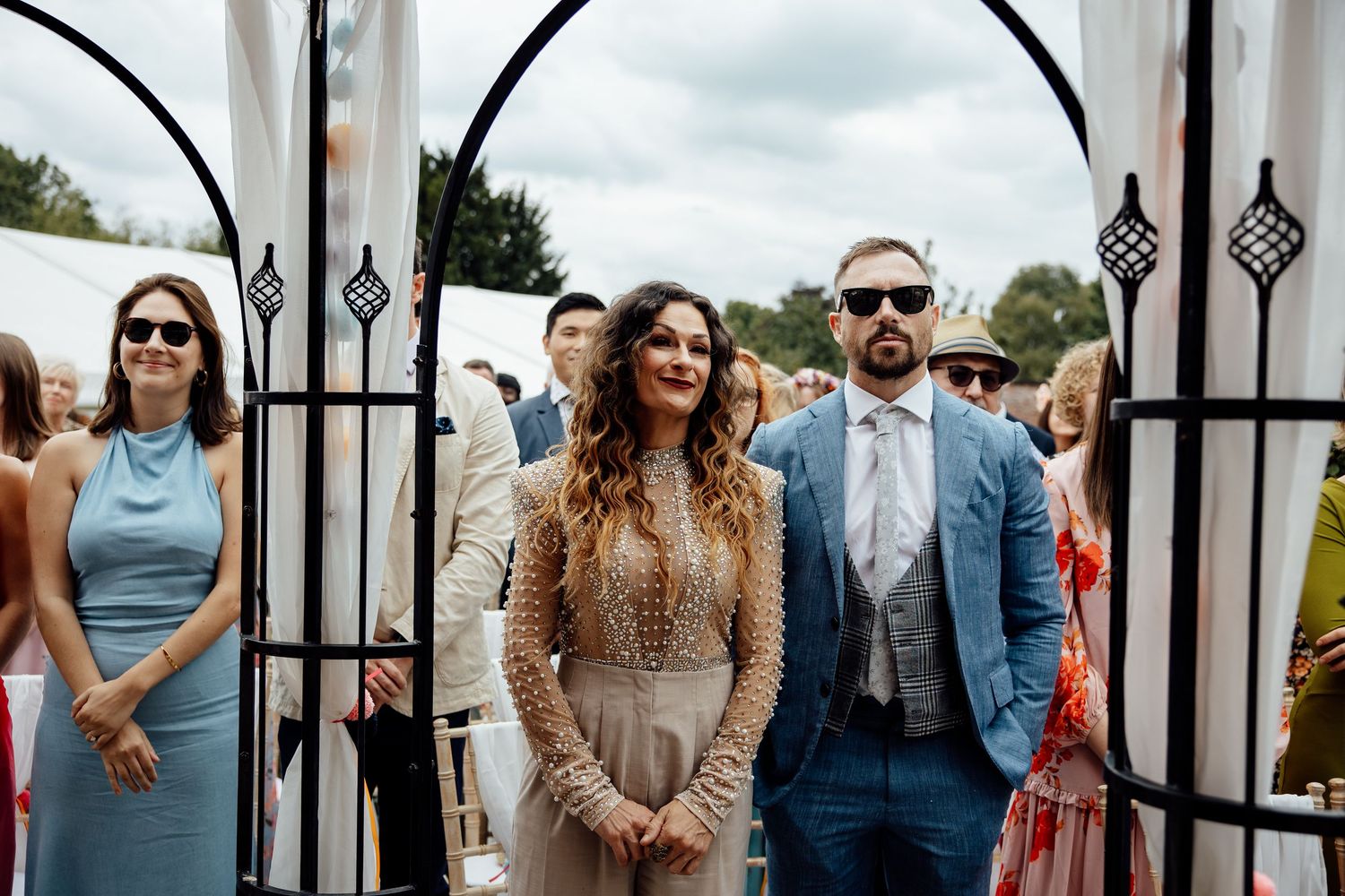 Elegantly dressed wedding guests stand beneath ornate iron gates at an outdoor ceremony.