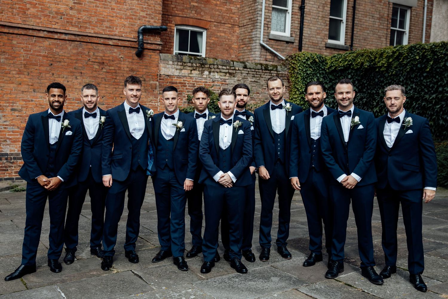 Groomsmen in dark suits pose together against brick building exterior.