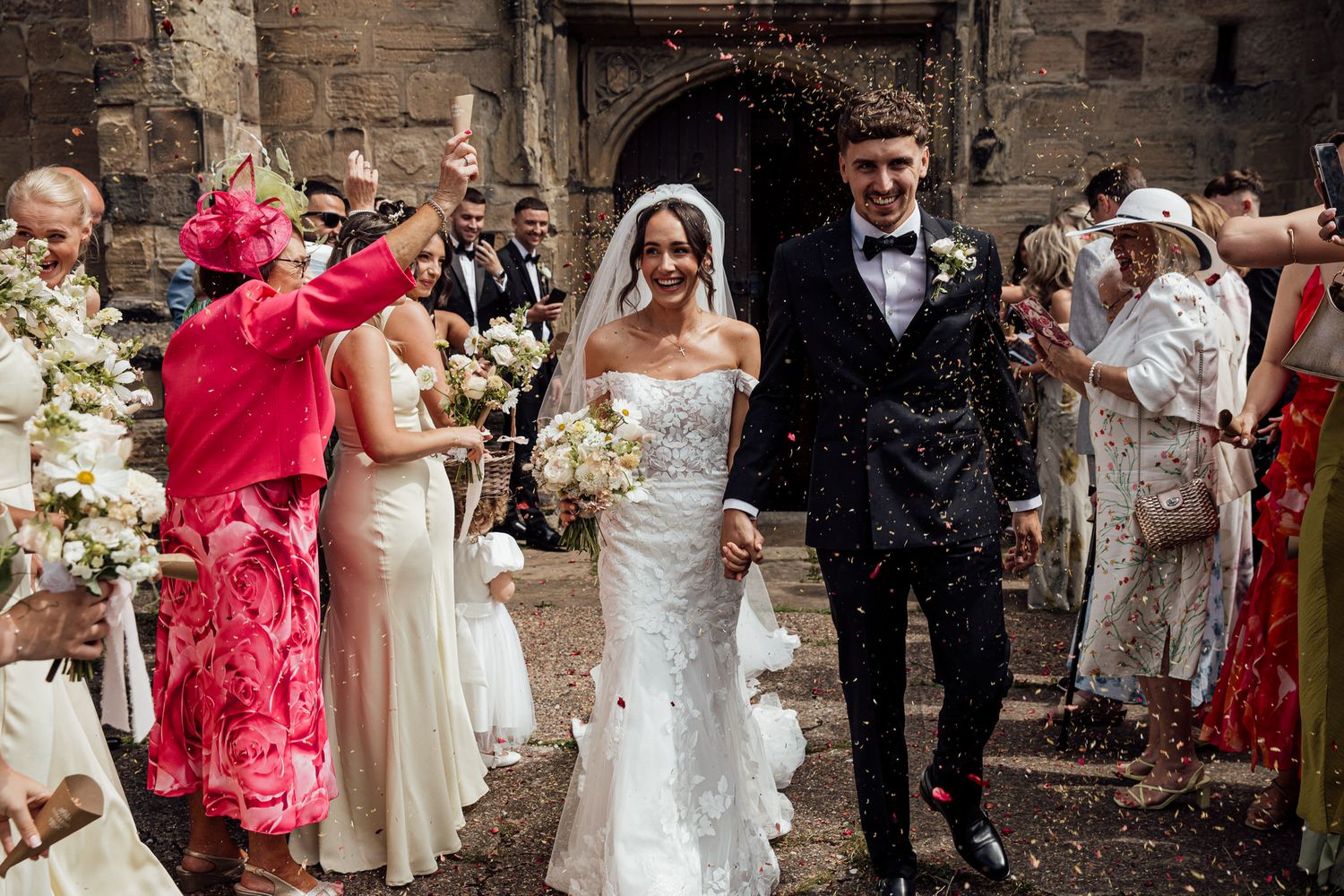 Wedding party celebrating with confetti and laughter outside historic stone building.