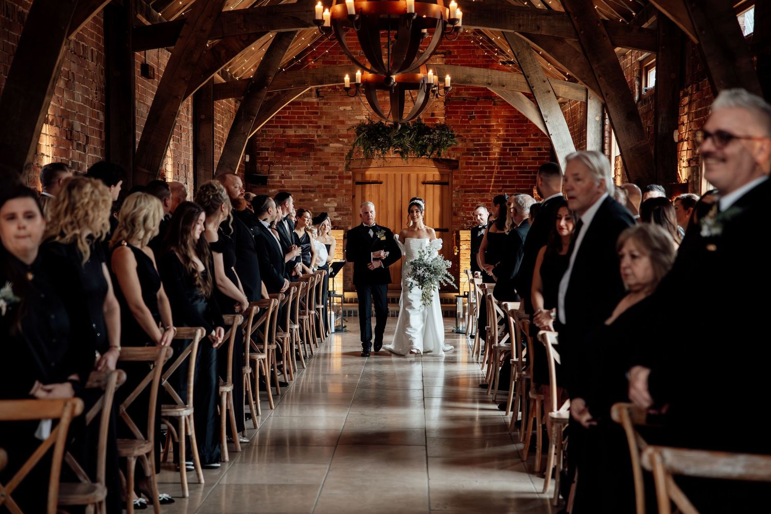 Wedding guests seated in elegant cross-back chairs inside historic brick venue with dramatic lighting.