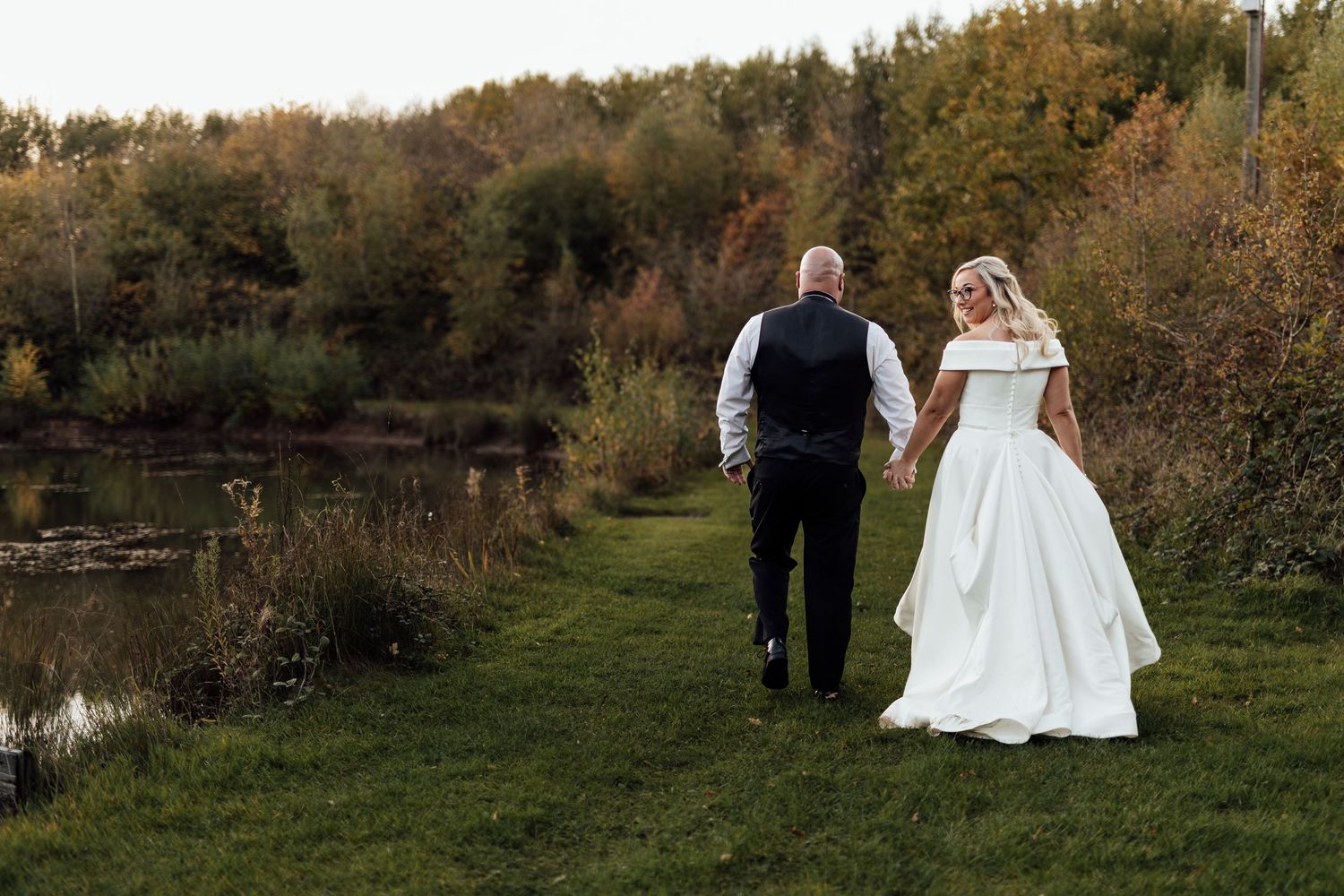 Newlyweds walk hand in hand along grassy path near pond during sunset.