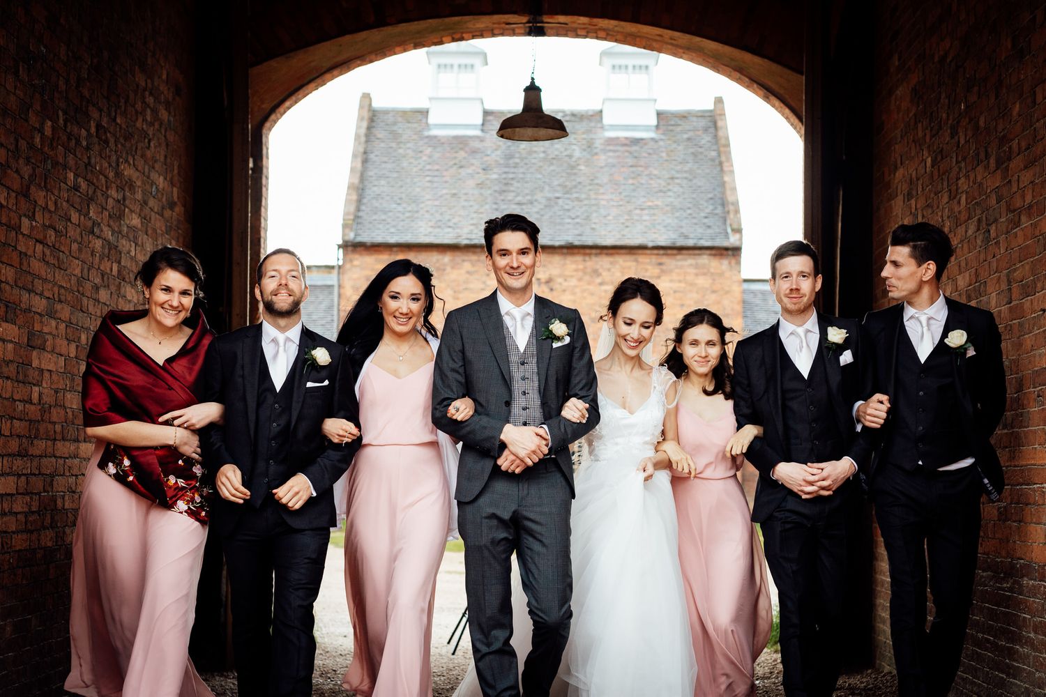 Wedding party lined up against rustic brick wall in formal attire.