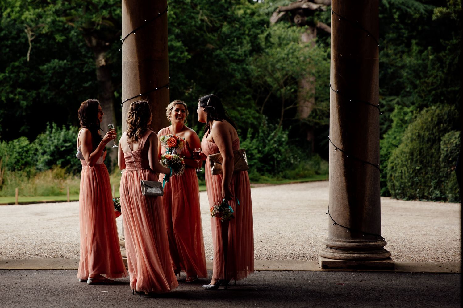 Bridesmaids in coral dresses stand together under stone colonnade.
