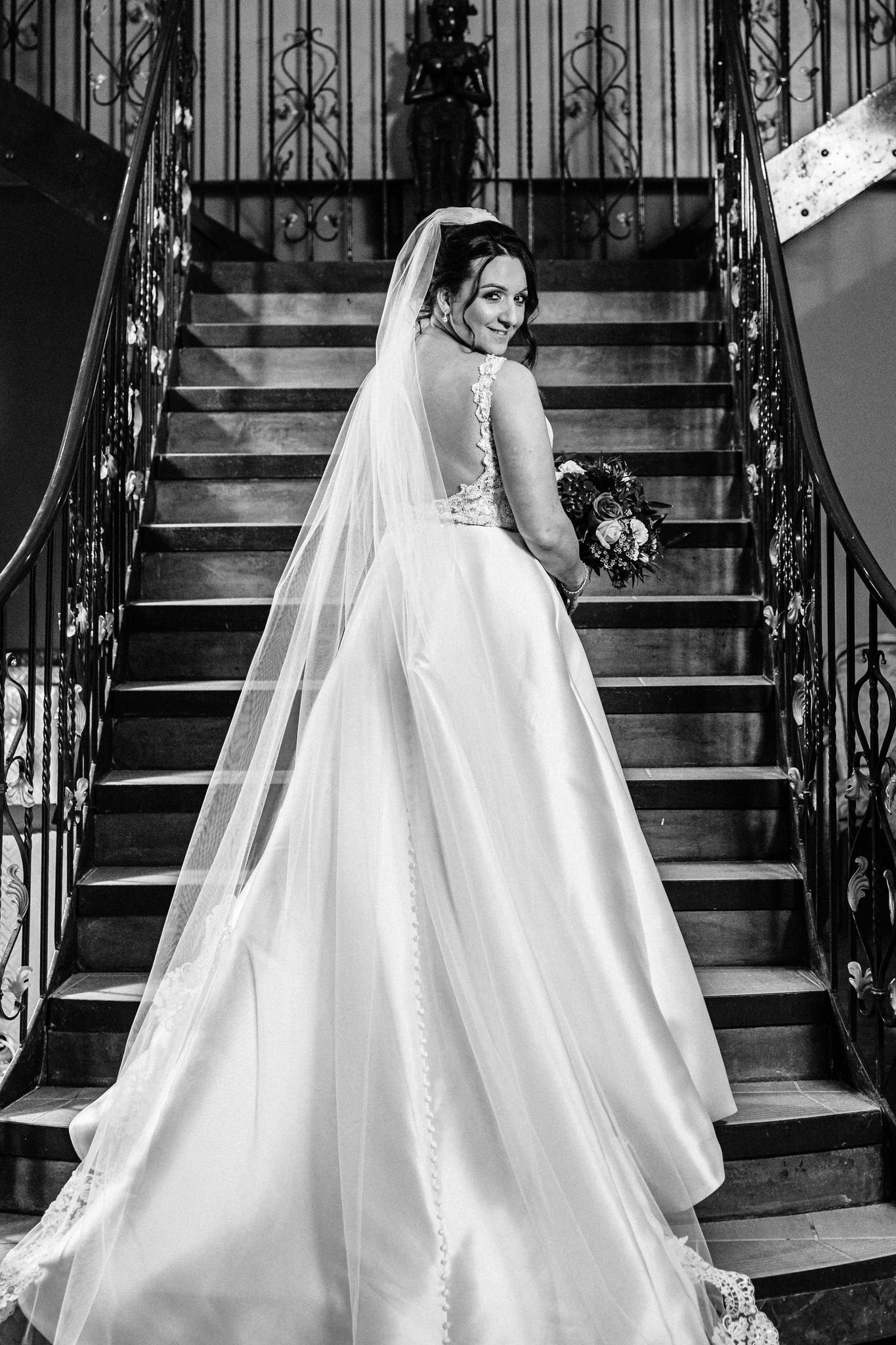 Flowing white wedding dress captured on elegant staircase with ornate iron railings.