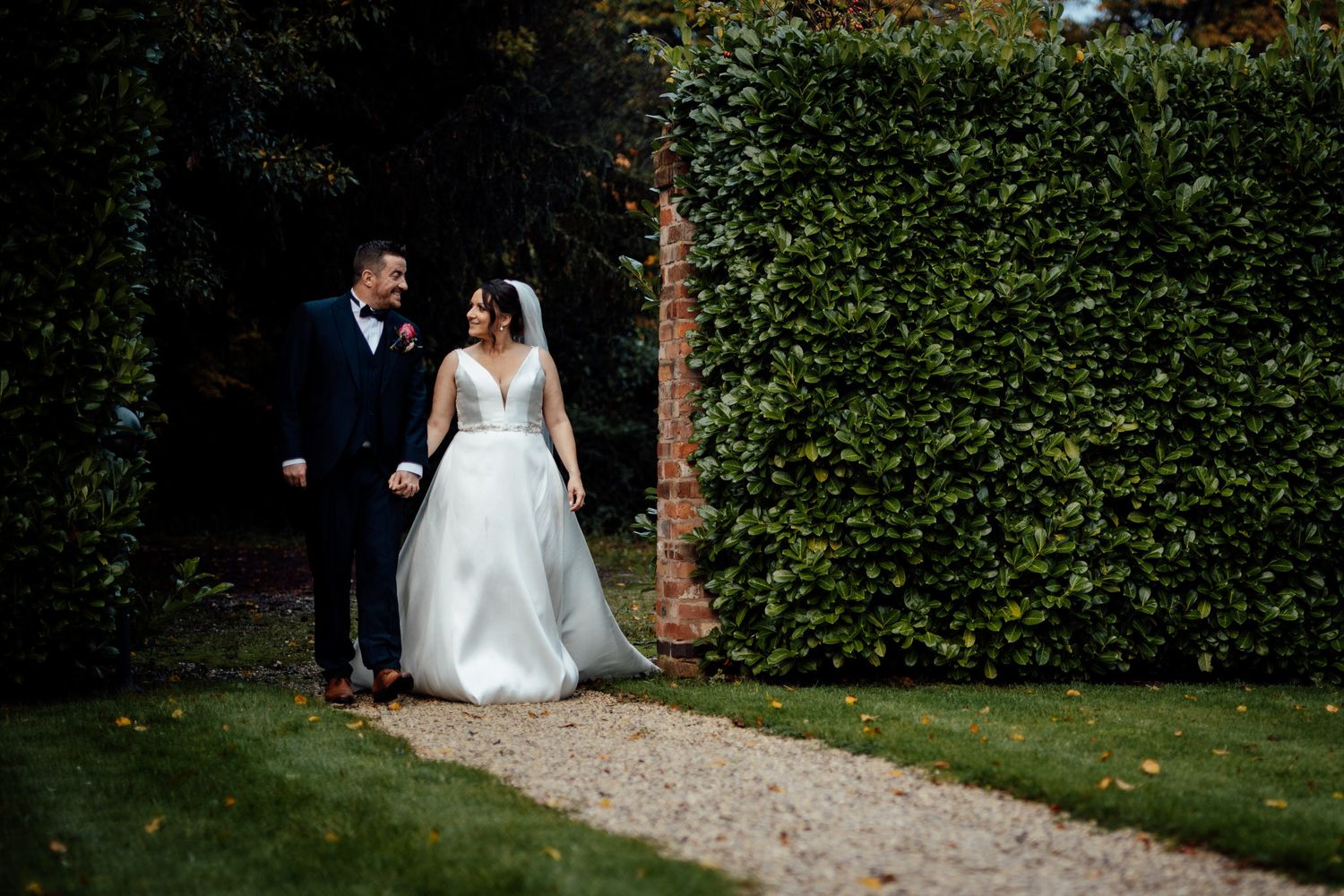 Wedding couple strolling through a garden path with lush green hedges.