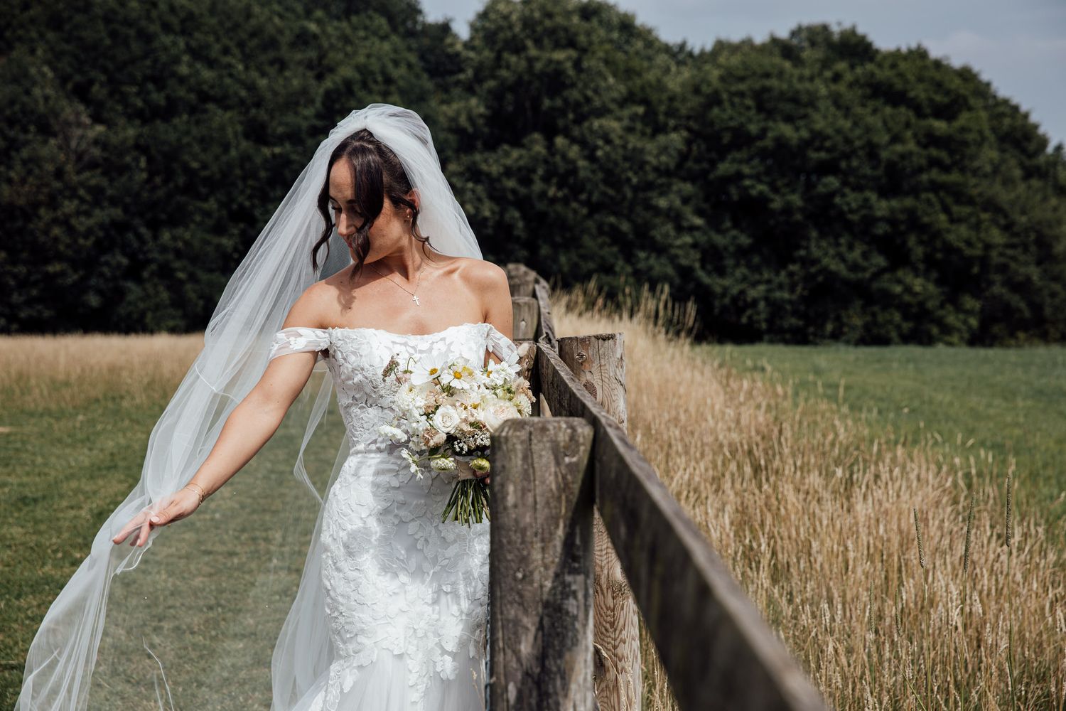 Bride in white dress poses near rustic wooden fence with scenic countryside backdrop.