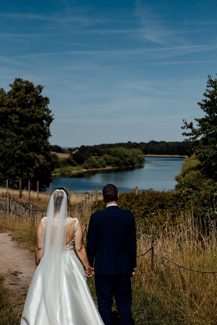 A couple walks along a scenic lakeside path surrounded by greenery on their wedding day.