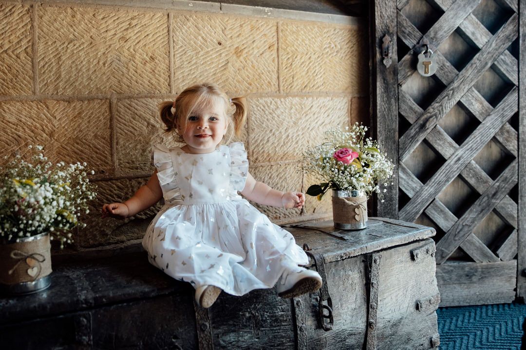 Child in white dress sitting on stone bench at wedding venue.