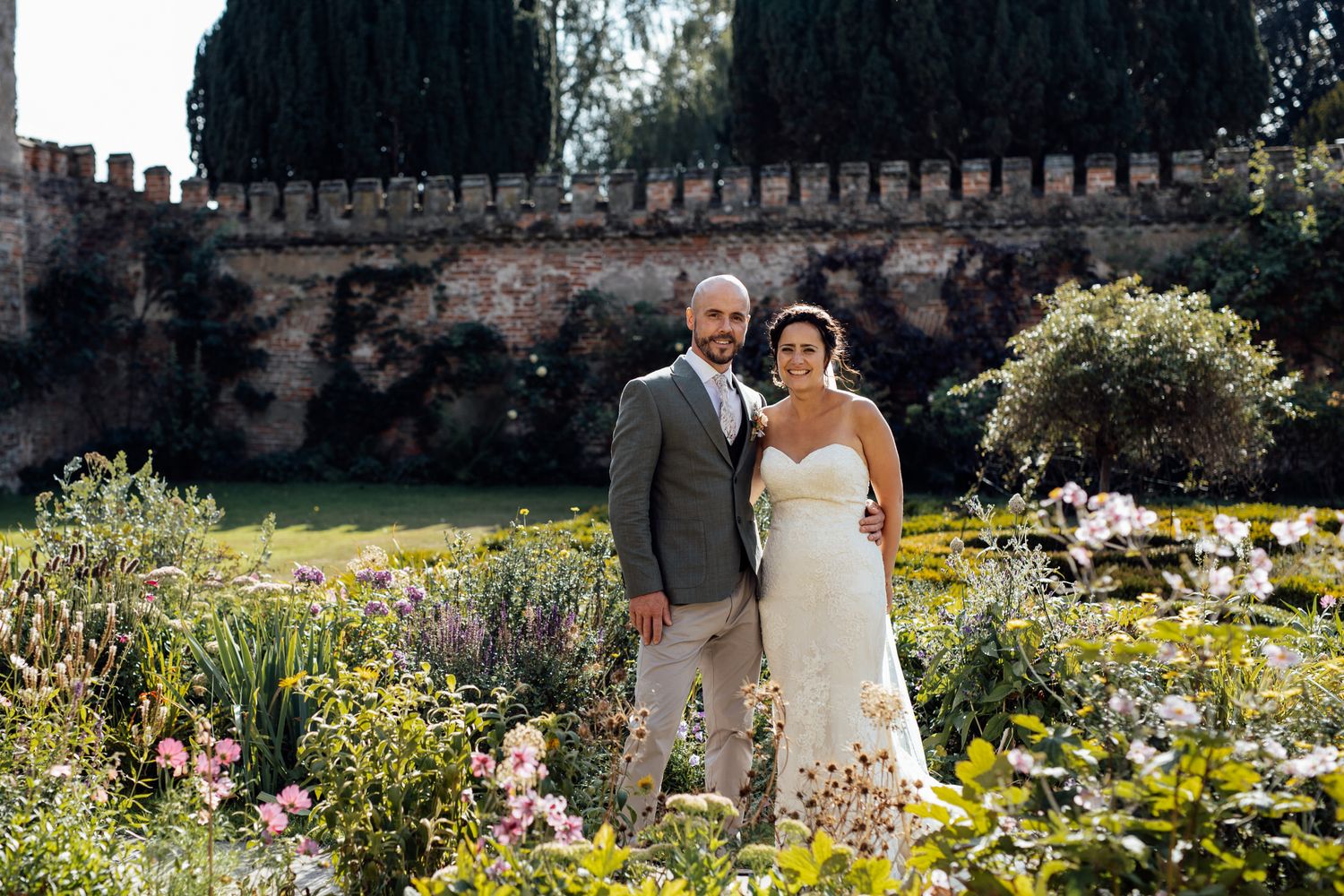 Newlyweds posing in blooming garden with stone wall backdrop.