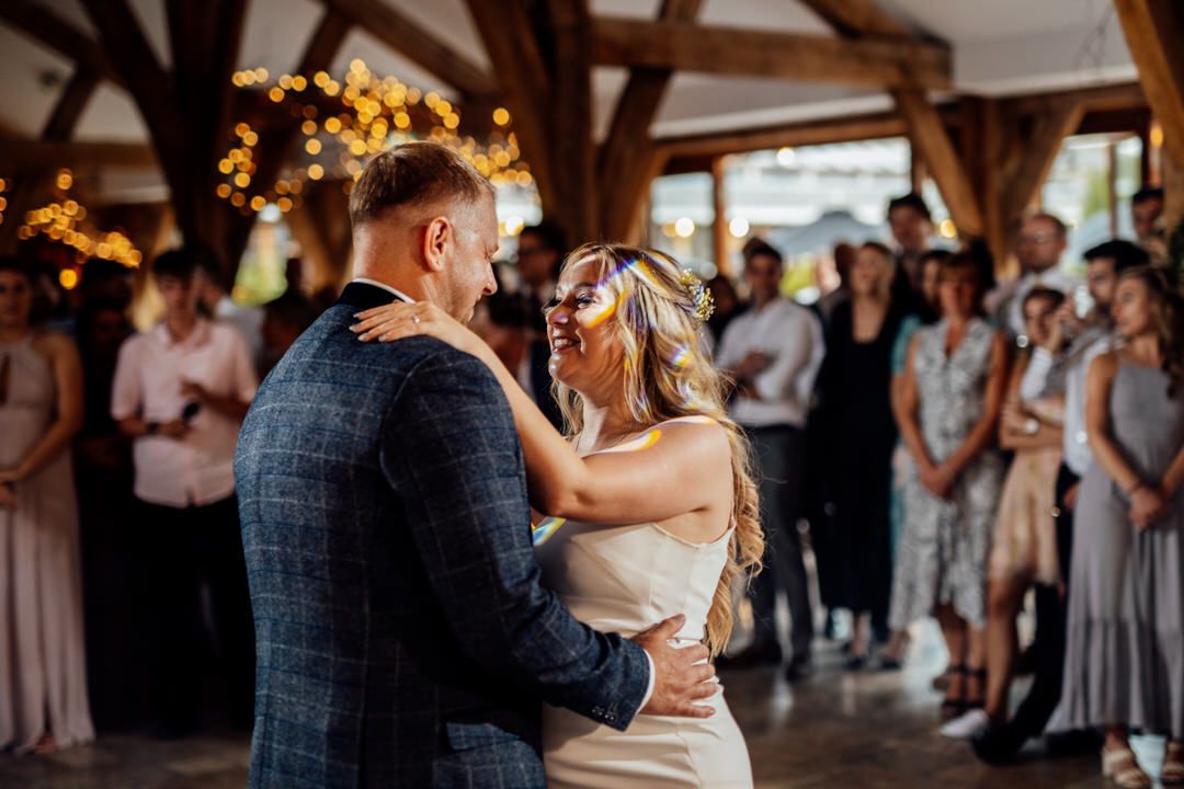 Wedding guests dance and celebrate in a rustic barn venue with exposed wooden beams and warm lighting.