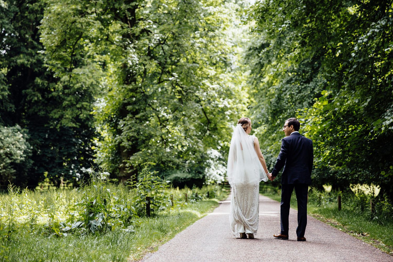A couple walks along a tree-lined path in a lush green garden setting on their wedding day.