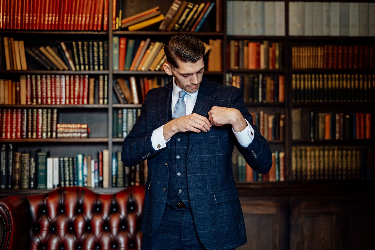 A person in a navy suit adjusts their tie while standing in front of a library bookshelf with leather-bound books.