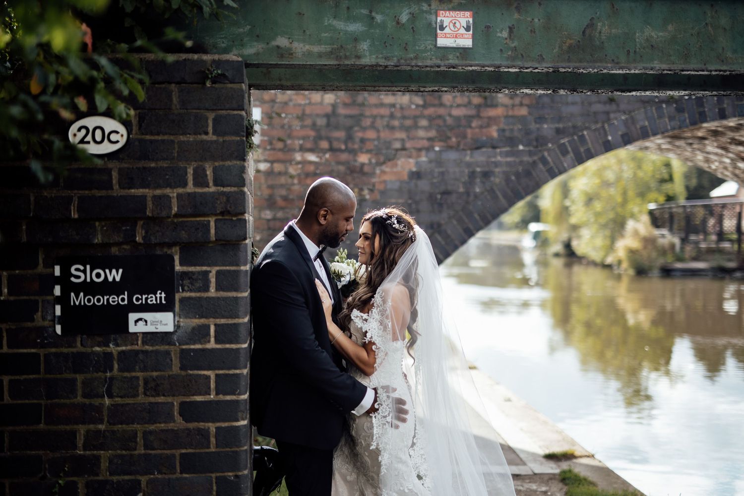 Newlyweds embracing near a brick wall with water reflection in background.