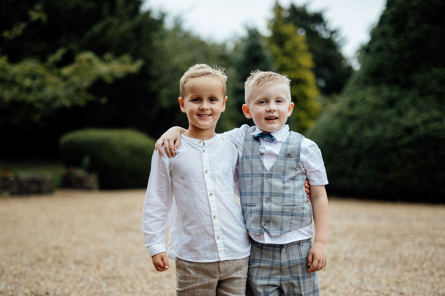 Two young boys in formal attire stand together outdoors against a natural backdrop.