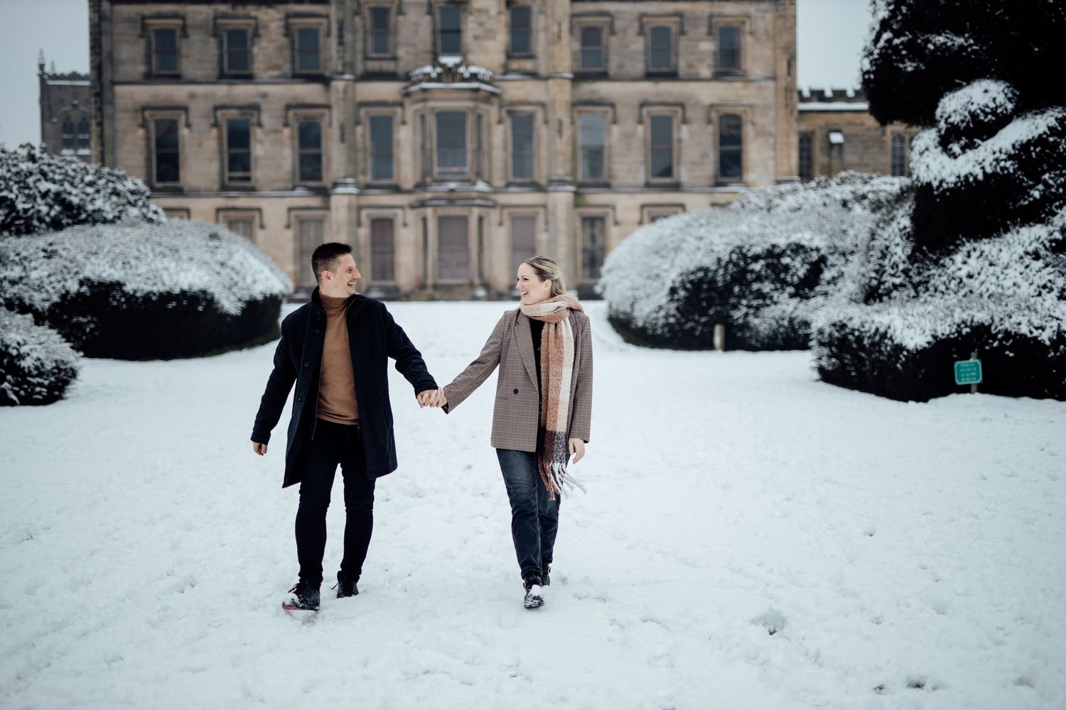 A romantic winter engagement photoshoot in front of a historic stone building covered in snow.