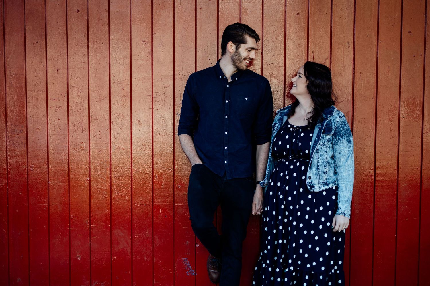 A couple poses against a rustic red wooden wall in casual attire.