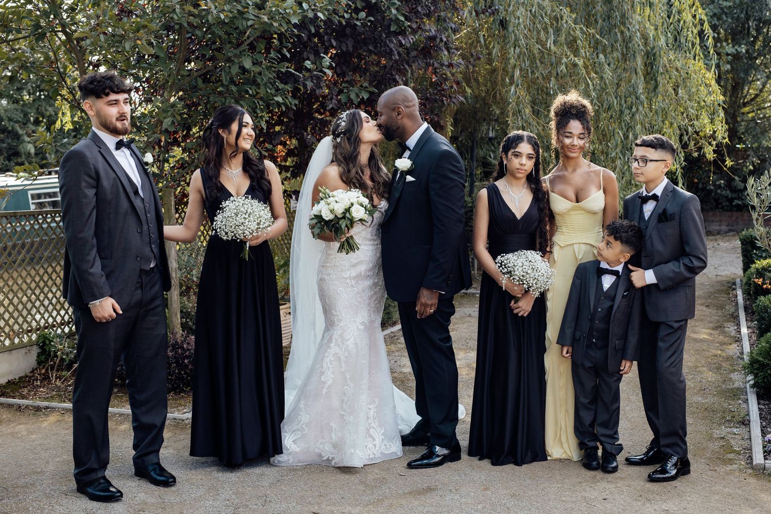 Wedding party in formal attire poses outdoors under willow trees at garden venue.