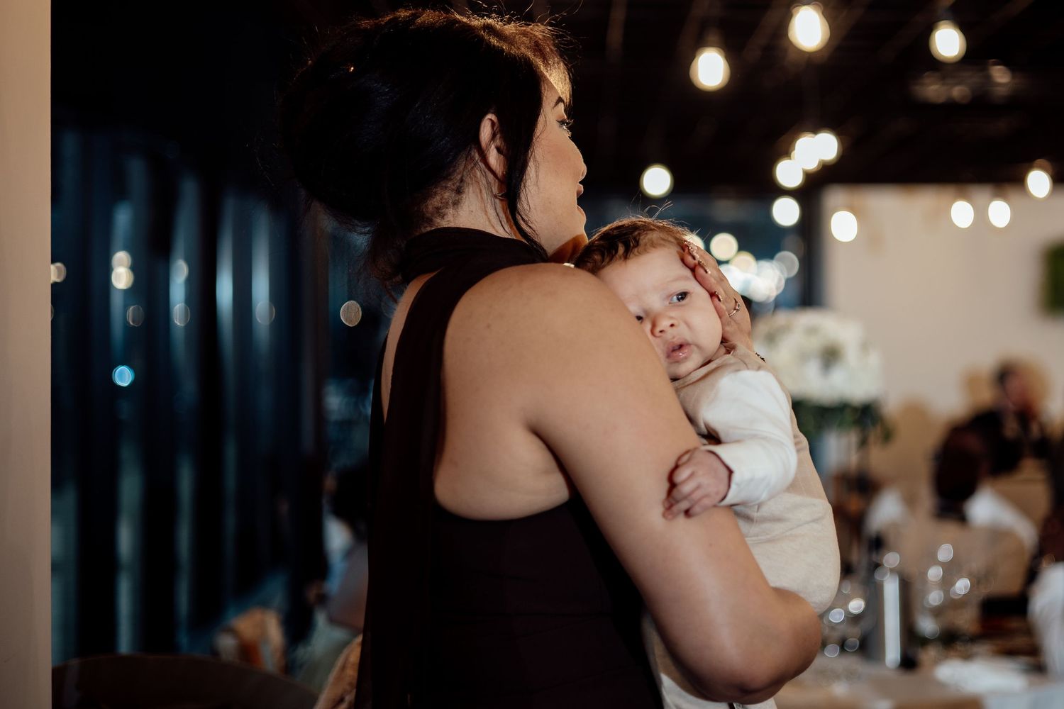 A woman gently holds a sleeping baby at a warmly lit wedding reception venue.