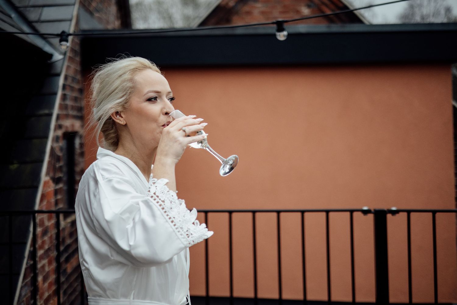 Blonde woman in white dress sipping champagne on an outdoor balcony.