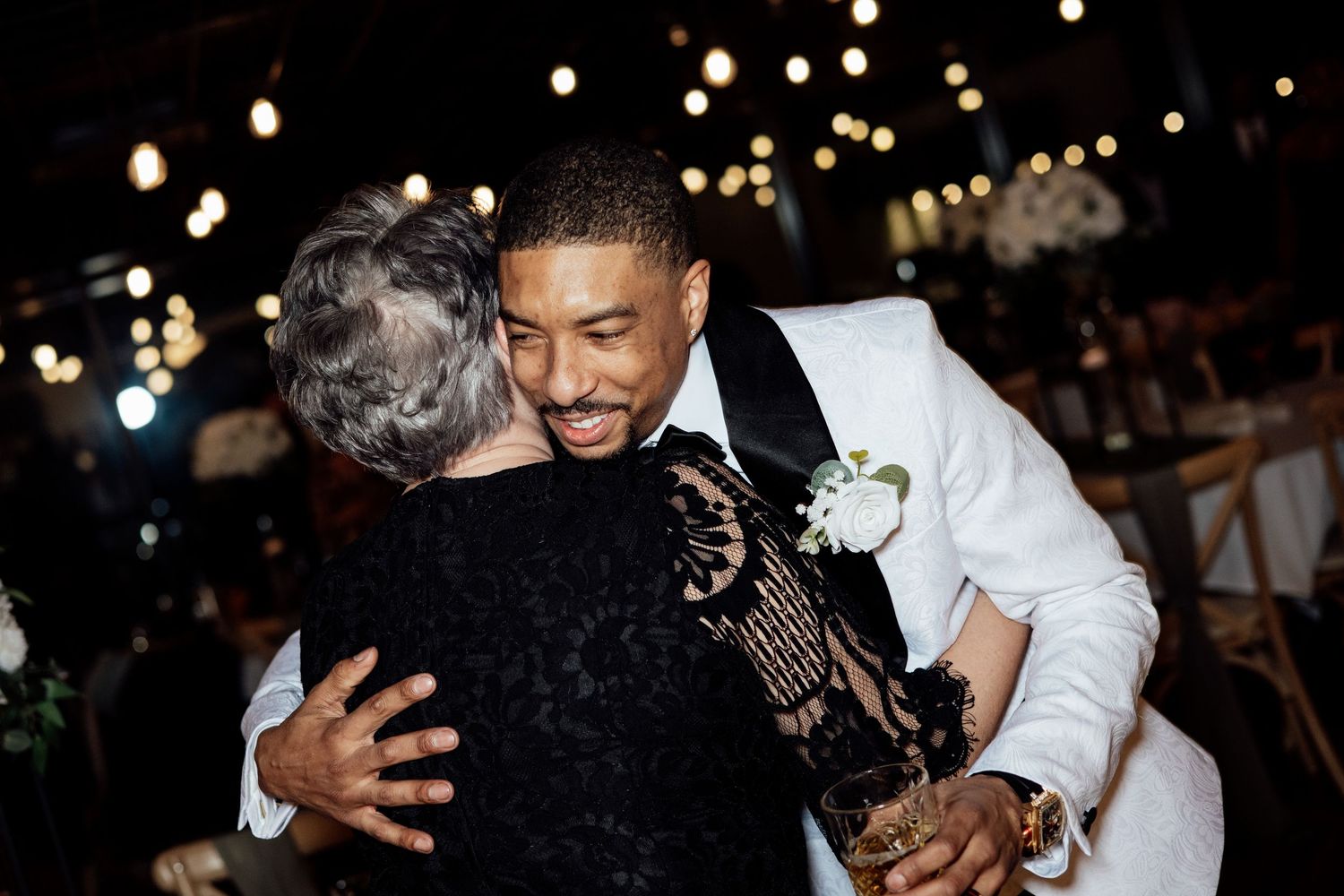 Groom in a white tuxedo shares a warm embrace with an elderly woman at the wedding reception.